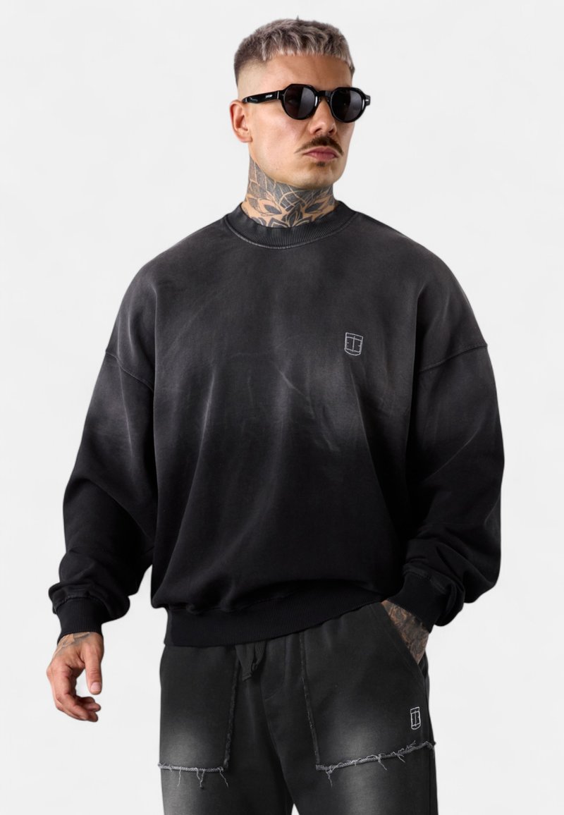 Schwarzes, übergroßes Sweatshirt mit Rundhalsausschnitt, das eine strukturierte Oberfläche und ein kleines Logo auf der Brust aufweist. Kombiniert mit ausgefransten schwarzen Hosen.