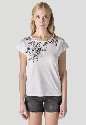 Mujer rubia con camiseta blanca de manga corta con estampado floral negro en el hombro y shorts vaqueros negros, de pie frente a un fondo gris.