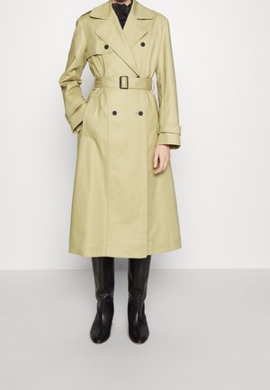 Femme portant un long trench-coat beige à double boutonnage avec une ceinture à la taille et des bottes noires montant jusqu'aux genoux, debout devant un fond uni.