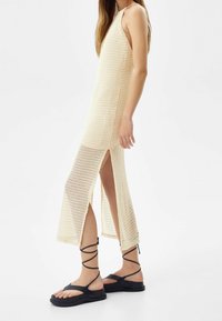 Beige pletené šaty s bočními rozparky, s designem na zavazování za krk. V páru s černými sandály na řemínky a strukturovaným povrchem.