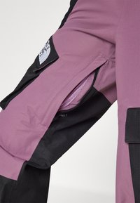 Lilla ja musta värvi õueriided, millel on lukuga võrgust taskud ja tekstureeritud kangas, ning reie peal on brodeeritud logo.