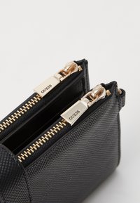 Borsa a tracolla nera con texture, dotata di due zip dorate, con il nome del brand impresso, e una forma strutturata con hardware minimale.