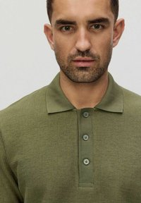 Zielona koszulka polo z prążkowanym wzorem, o fakturze, klasycznym kołnierzyku i zapięciu na trzy guziki. Posiada jasnoszare guziki i luźny krój.