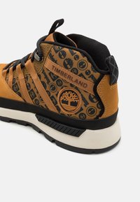 Braune und schwarze Mid-Top Sneaker mit einem gemusterten Obermaterial aus Stoff, Gummisohle und eingeprägtem Timberland-Logo an der Seite.