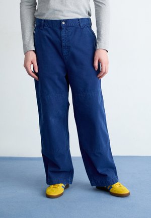 Person iført blå bukser med vide ben, grå langærmet skjorte og gule sneakers, stående på blåt tæppe mod en hvid væg.