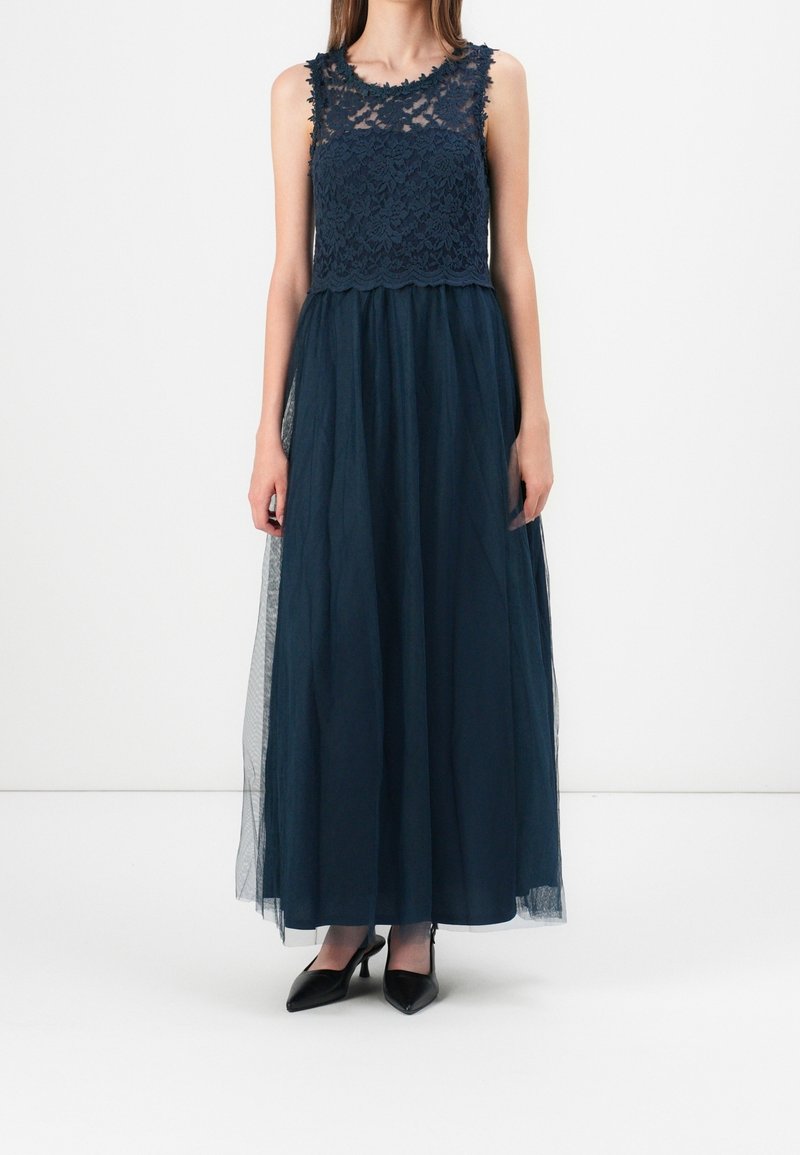 Femme portant un haut en dentelle bleu foncé sans manches et une longue jupe transparente, associée à des talons noirs pointus, debout devant un fond blanc.
