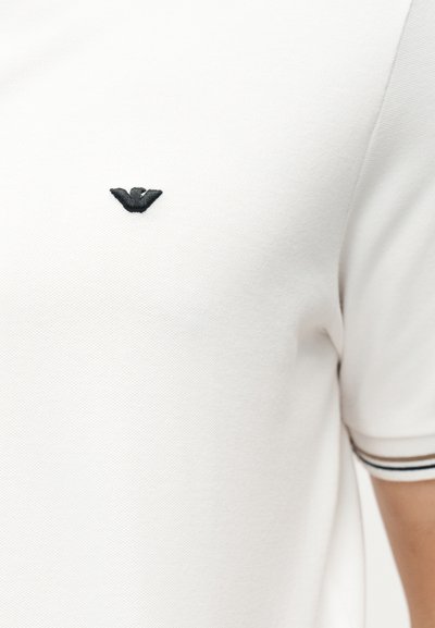 Λευκό πολο με κοντά μανίκια και υφή, με κεντητό μαύρο λογότυπο αετού στο στήθος και μαύρη και λευκή ριγωτή επένδυση στην άκρη του μανικιού.