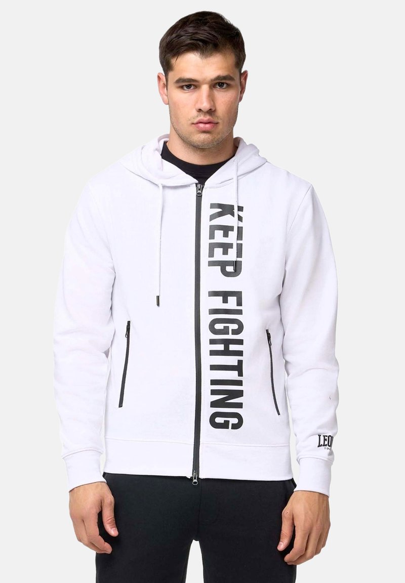 Homme portant un sweat à capuche blanc zippé avec un texte vertical noir « KEEP FIGHTING » et un pantalon noir, debout face à un fond uni.