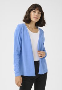 Gilet bleu clair en tissu doux, design à ouverture sur le devant, manches longues et poignets côtelés. Porté par-dessus un top blanc et associé à un pantalon noir.