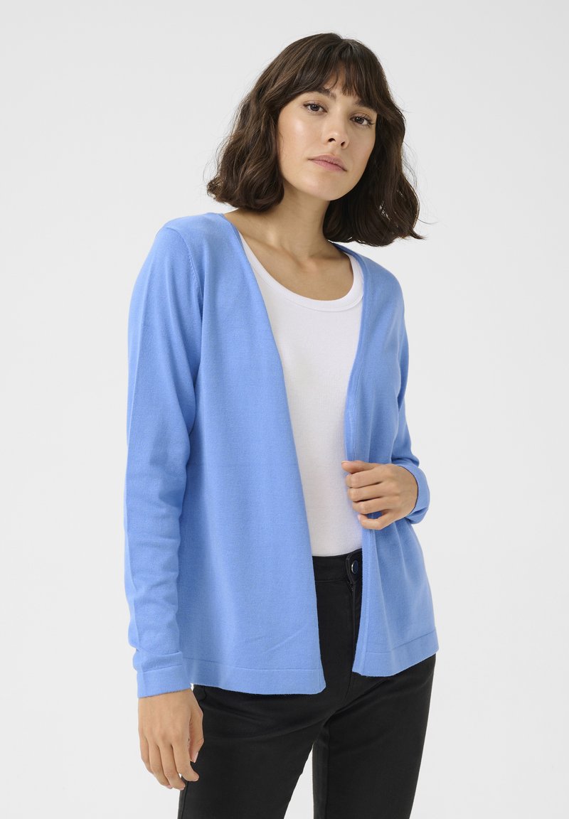 Gilet bleu clair en tissu doux, design à ouverture sur le devant, manches longues et poignets côtelés. Porté par-dessus un top blanc et associé à un pantalon noir.