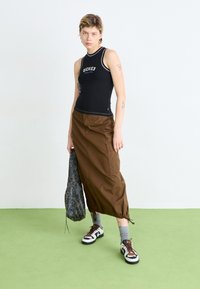Svart ermeløs topp med "Dickies"-logo, brun cargo-lignende skjørt, mønstret snorveske, og svart-hvite sneakers med grå sokker.