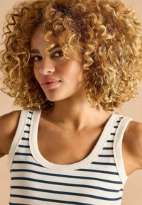 Striped tank top in white with navy blue stripes, featuring a round neckline and a ribbed texture. Model has curly blonde hair.