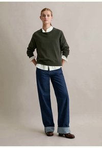 Junge Frau steht mit den Händen in den Taschen, trägt einen dunkelgrünen Pullover über einem weißen Hemd, weit geschnittene, gekrempelte Jeans und braune Schuhe.