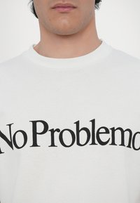 Hvit bomull t-skjorte med setningen "No Problemo" i fet svart skrift. Designet er enkelt med en klassisk rund hals.