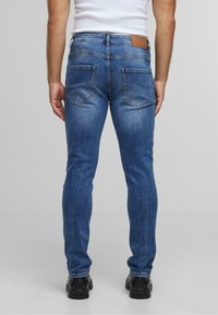 Jeans de denim azul con corte slim, que presentan un parche de cuero marrón en la cintura y dos bolsillos traseros. La tela tiene una apariencia de lavado medio.