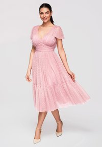Femme souriante, portant une robe midi rose à pois avec des manches volantes et des talons beige à bride à la cheville, debout sur un fond gris clair.