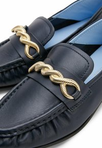 Mocassini in pelle blu navy con un accento in catena dorata attorcigliata, caratterizzati da una punta rotonda e dettagli arricciati lungo i bordi. Interno azzurro chiaro.