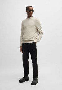 Maglione strutturato beige chiaro, pantaloni cargo neri con tasche e stivali neri con lacci. Occhiali da sole scuri completano l'outfit.