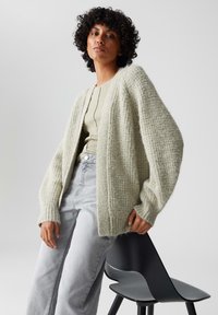 Lichtgroene, getextureerde gebreide cardigan met een ontspannen pasvorm en verlaagde schouders, gedragen over een beigetop en gecombineerd met lichtgrijze jeans.