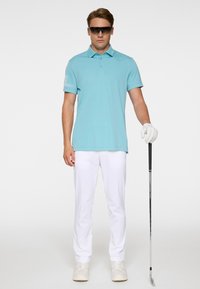 Homme portant un polo bleu clair, un pantalon blanc, un gant de golf et des lunettes de soleil, tenant un club de golf, debout devant un fond blanc.