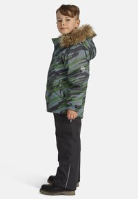 Roheline ja musta mustriga talvejope karusnahaga voodriga kapuutsiga, seljas mustad püksid ja robustsed mustad saapad, seisvas poosis.