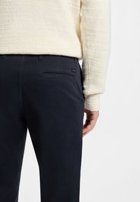 Temno modre bombažne hlače s tesnim krojem, ki imajo zadnji žep in subtilno zastavico z logotipom. Nošene s teksturiranim kremnim džemperjem iz pletenine.