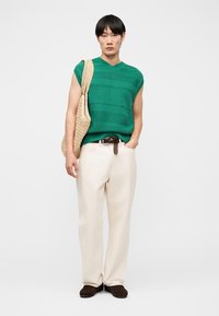 Jeune personne portant un haut sans manches en maille verte, un pantalon large crème, des chaussures marron, et un sac fourre-tout tissé beige porté sur une épaule.