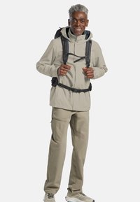Licht beige waterdichte jas met verstelbare kap, voorritsen en meerdere zakken, gecombineerd met bijpassende beige broek en wandelschoenen.