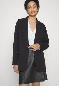 Blazer noir oversize avec revers en notch, fermeture à un bouton et doublure intérieure bleue, associé à un blouse blanche et une jupe en cuir noir.