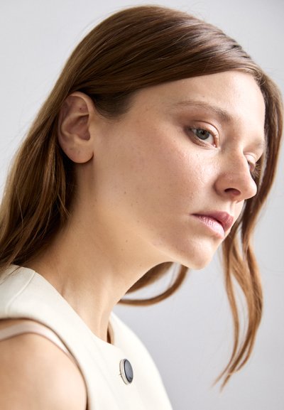 Vue de profil d'une personne avec des cheveux raides châtain clair et un maquillage neutre. Elle porte un haut sans manches de couleur crème avec un accent de bouton noir.