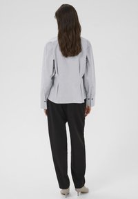 Kvinde med langt brunt hår iklædt en lys grå langærmet bluse, sorte løstsiddende bukser og lysegrå hæle, som står vendt væk.