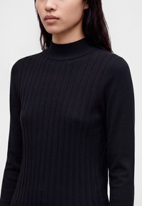 Femme portant un pull noir côtelé à manches longues avec un col montant, montrée des épaules jusqu'au milieu du torse sur un fond uni.