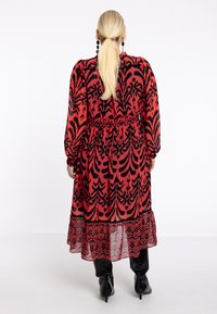 Langes rotes Kleid mit schwarzem Blumenmuster, elastischem Bund, langen Ärmeln und geschichteter, transparenter Rüsche am Saum. Getragen mit knielangen schwarzen Stiefeln.