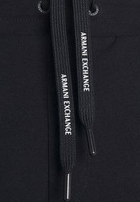 Cuerda negra con estampado en blanco "ARMANI EXCHANGE", unida a una tela negra lisa con textura textil y ojales reforzados.