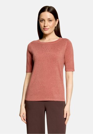 Vrouw met stijl donker haar die een kort mouw gebreide top in rozekleur en donkerbruine broek draagt, staand tegen een effen lichte achtergrond.