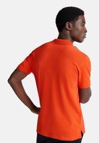 Muž v jasně oranžovém polokošili s krátkým rukávem, stojící bokem s jednou rukou na bok, před jednobarevným bílým pozadím.