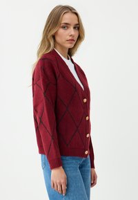 Młoda kobieta w czerwonym kardiganie na guziki z czarnym wzorem w romby, nosząca biały top i niebieskie dżinsy, stojąca bokiem na białym tle.