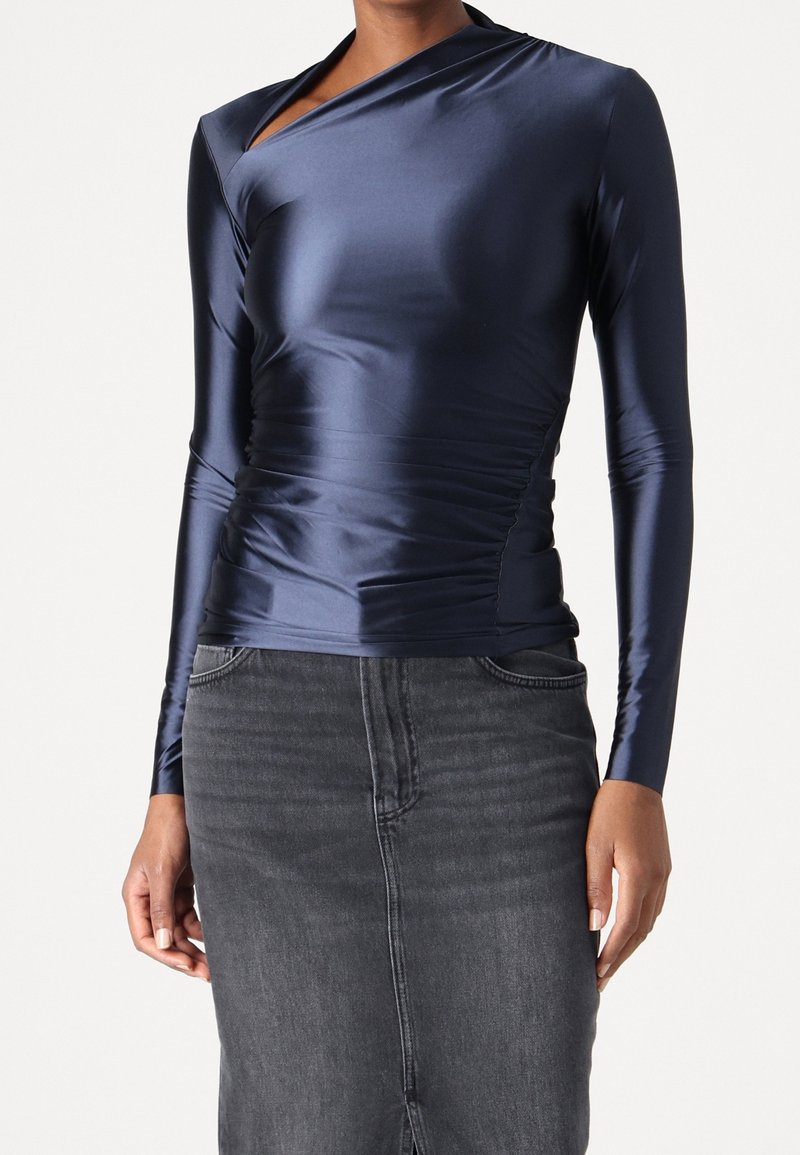 Marineblå langærmet top i glat, skinnende stof med asymmetrisk halsudskæring og rynkede sider, parret med en tætsiddende denimnederdel.