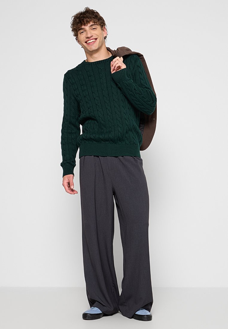Suéter de punto en verde oscuro, pantalones de pierna ancha grises y zapatos azules. La modelo está de pie con una chaqueta marrón caída sobre un hombro.