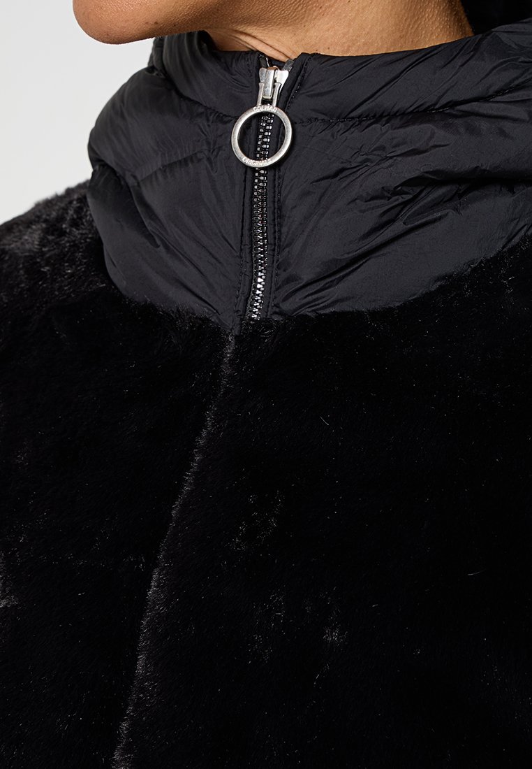 Primo piano di una giacca nera con texture in pelliccia morbida e colletto alto in nylon, dotata di una cerniera con anello metallico argento sul collo.
