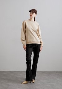 Béžová mikina s kulatým výstřihem, černé cargo kalhoty s bočními kapsami, hnědá kšiltovka a krémové muly. Hladká textura, ležérní design.