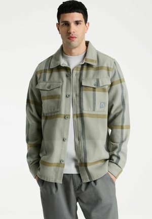 Jonge man draagt een groen-beige geruit jasje met knopen over een wit overhemd met grijze broek, staand met handen in de zakken tegen een witte achtergrond.
