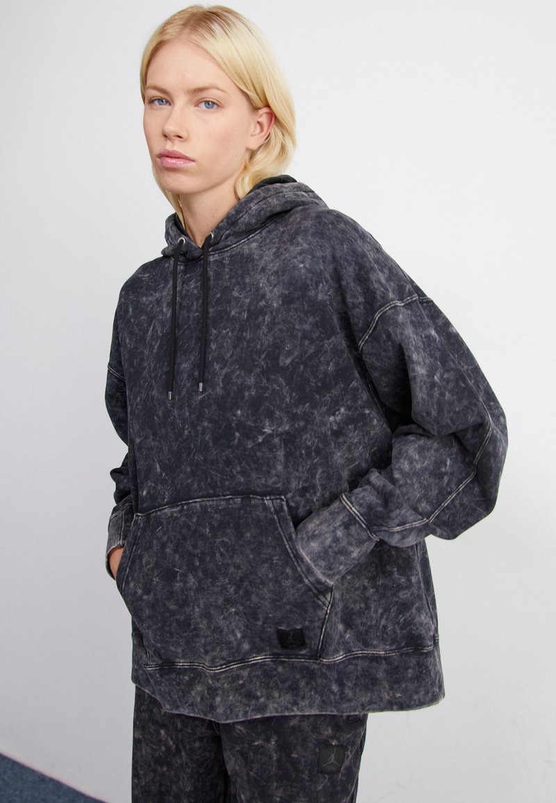 Sudadera con capucha en un lavado negro desgastado, con un bolsillo frontal, capucha con cordón y un corte relajado. La tela tiene un aspecto suave y texturizado.