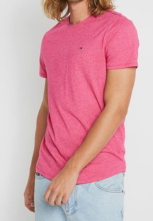 Man met schouderlang haar, gekleed in een roze T-shirt met korte mouwen en een ronde hals, en lichtblauwe spijkerbroek, staand tegen een witte achtergrond.