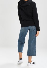 Person som bär en svart hoodie, avklippta blå denimjeans med slitna kanter och vita Adidas-sneakers med svarta ränder, står på vit golv.