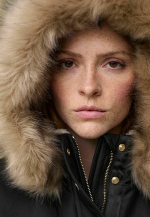Parka noire avec une capuche en fourrure, dotée d'une fermeture éclair dorée et de boutons-pression. Le modèle a la peau tacheté de rousseur et des yeux marron.