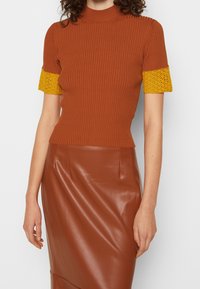 Ruskea, kammattua pintaa omaava toppi, jossa on korkea kaulus ja keltaiset virkatut lyhyet hihat, yhdistettyinä ruskeaan nahkainen kynähameeseen.