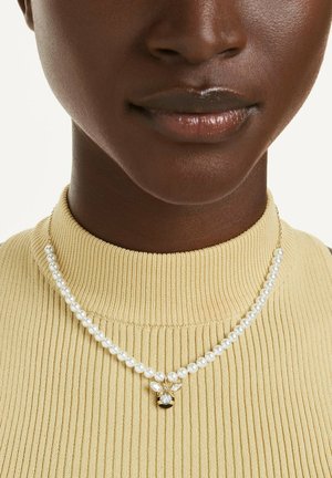 Close-up of a person wearing a pearl necklace with a gold bee pendant, paired with a light yellow ribbed turtleneck sweater.