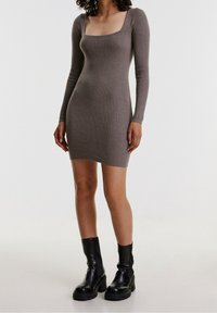 Kvinna klädd i en figursydd, långärmad, ribbad miniklänning i taupe med fyrkantig halsringning och svarta klumpiga ankelstövlar.