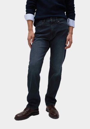 Uomo che indossa jeans blu scuro, un maglione blu navy con polsini di camicia azzurri e scarpe eleganti in pelle marrone, in piedi davanti a uno sfondo neutro.